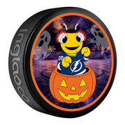 A black hockey puck featuring a circular Halloween-themed graphic. The graphic shows a cartoon bee wearing a Tampa Bay Lightning jersey sitting in a jack-o'-lantern, with a purple spooky background containing ghosts and bats.